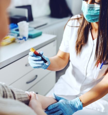 Female lab assistant in uniform, with protective mask and rubber gloves holding test tube with blood and holding patient's arm. Patient holding cotton on vein.